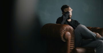 a-man-holds-his-head-while-sitting-on-a-sofa.jpg a man holds his head while sitting on a sofa